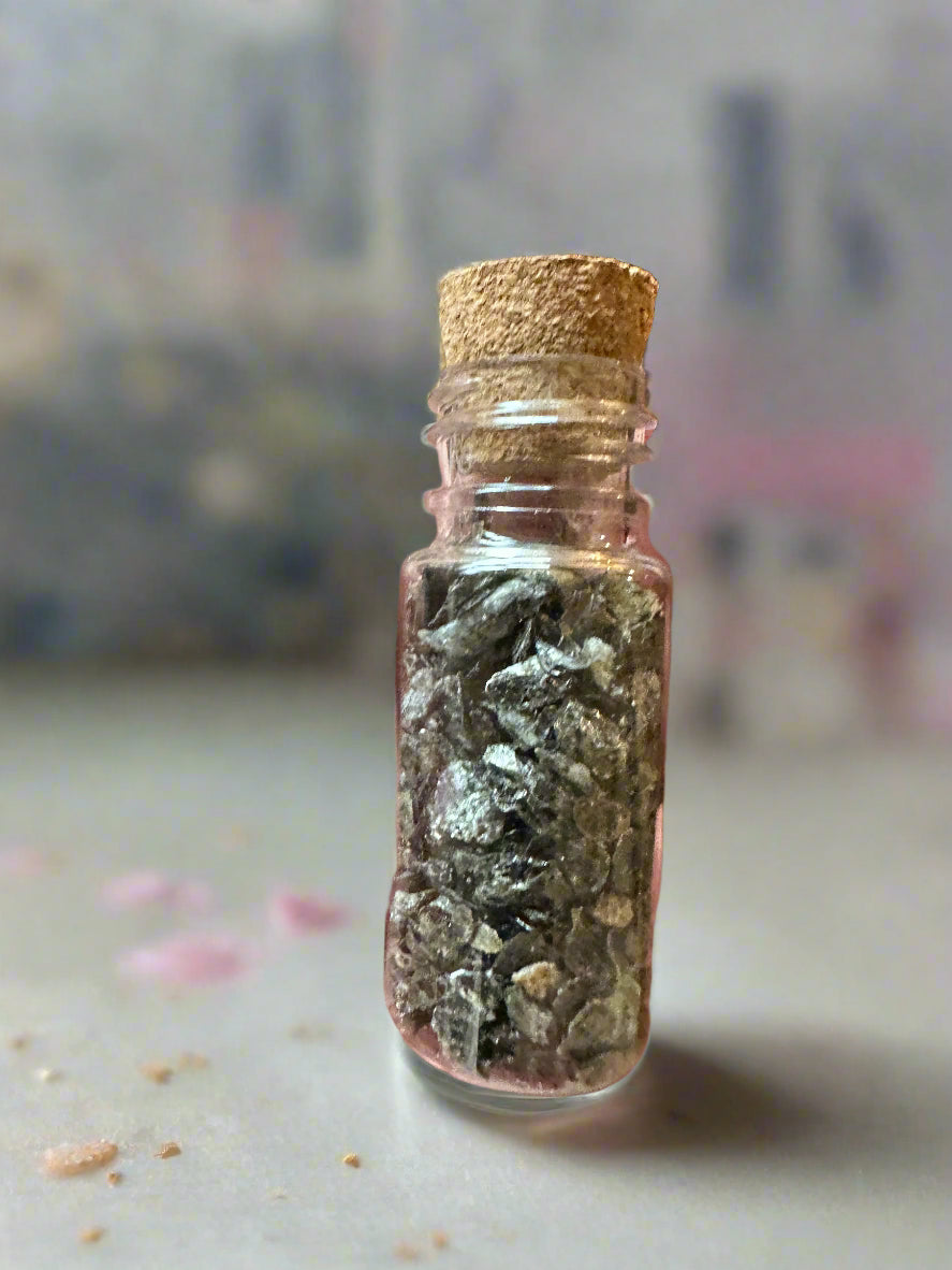 Hand holding a small glass bottle with cork lid containing green crystals against a blurred indoor background.